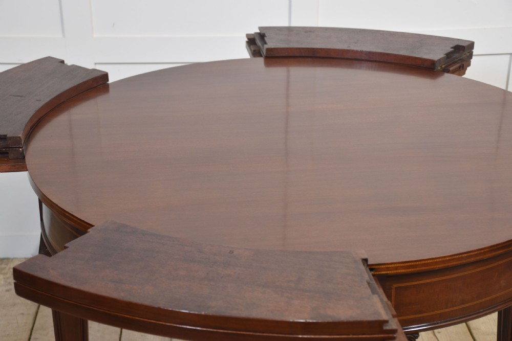 A dark mahogany circular dining table featuring outer leaf extension segments connected with brass hinges and supported by four fluted legs. View XII