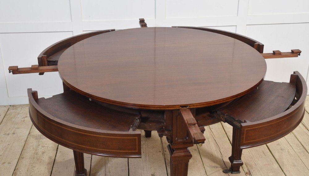 A dark mahogany circular dining table featuring outer leaf extension segments connected with brass hinges and supported by four fluted legs. View XIII