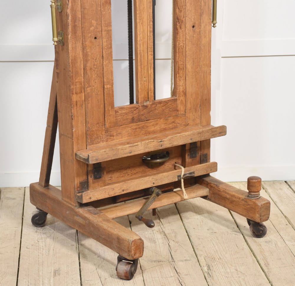 A large antique wooden artist studio easel with a crank handle mechanism standing on a pale wooden floor. View III