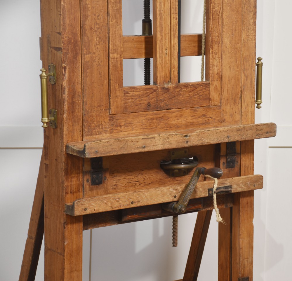 A large antique wooden artist studio easel with a crank handle mechanism standing on a pale wooden floor. View VIII