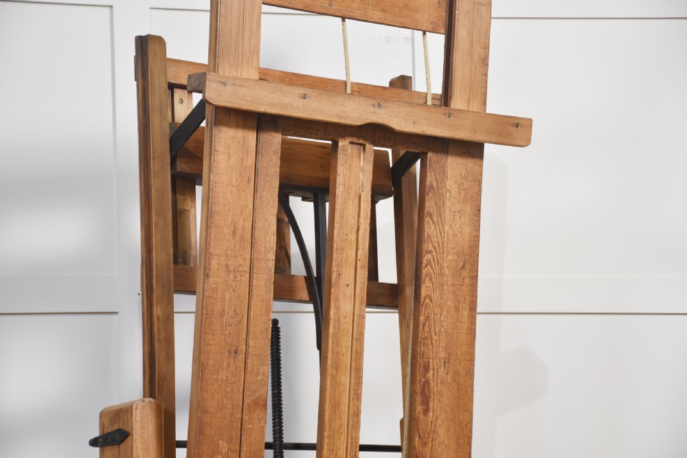 A large antique wooden artist studio easel with a crank handle mechanism standing on a pale wooden floor. View XV
