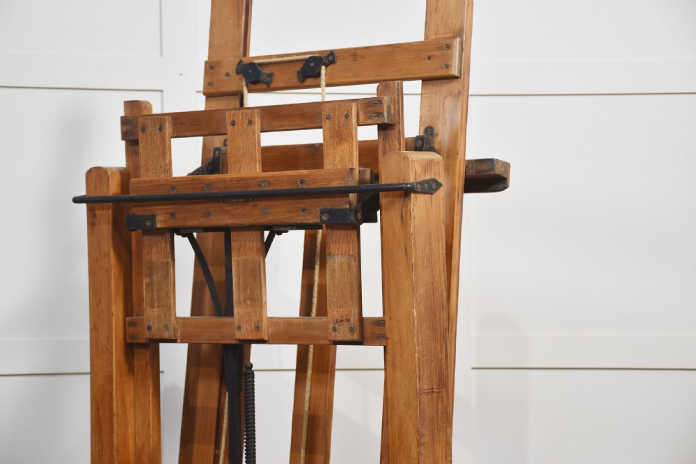 A large antique wooden artist studio easel with a crank handle mechanism standing on a pale wooden floor. View XIX