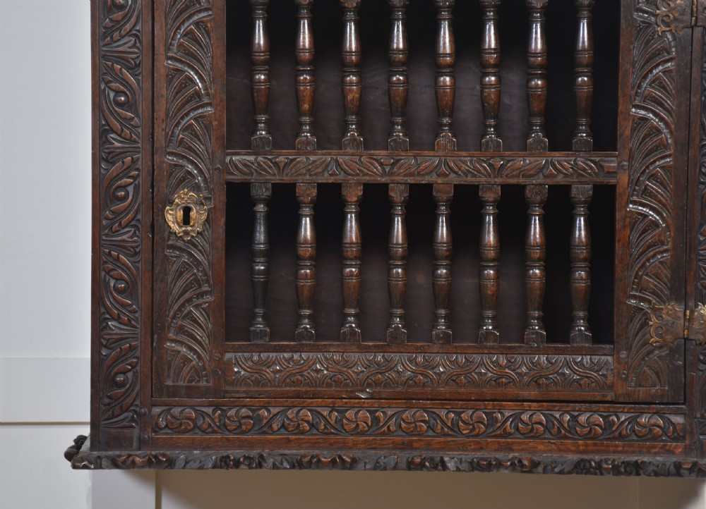 A wall-mounted 17th-century dark oak mural livery cupboard featuring hand-carved borders, turned wooden spindles, and brass hardware. View V