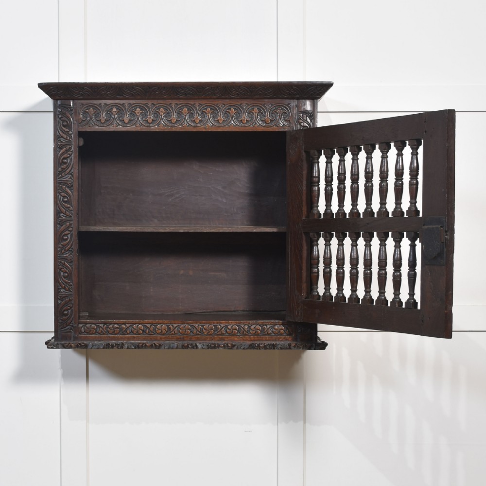 A wall-mounted 17th-century dark oak mural livery cupboard featuring hand-carved borders, turned wooden spindles, and brass hardware. View VI