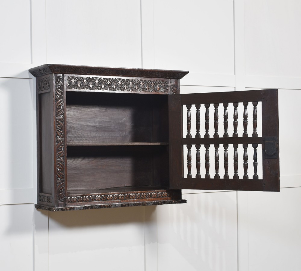 A wall-mounted 17th-century dark oak mural livery cupboard featuring hand-carved borders, turned wooden spindles, and brass hardware. View VIII