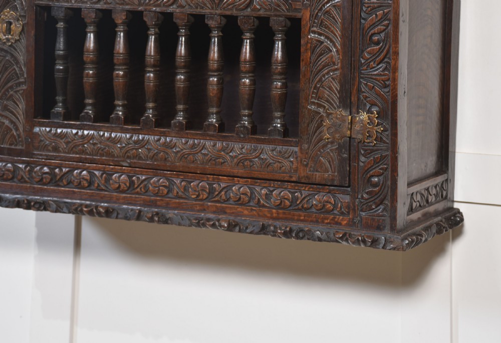 A wall-mounted 17th-century dark oak mural livery cupboard featuring hand-carved borders, turned wooden spindles, and brass hardware. View X