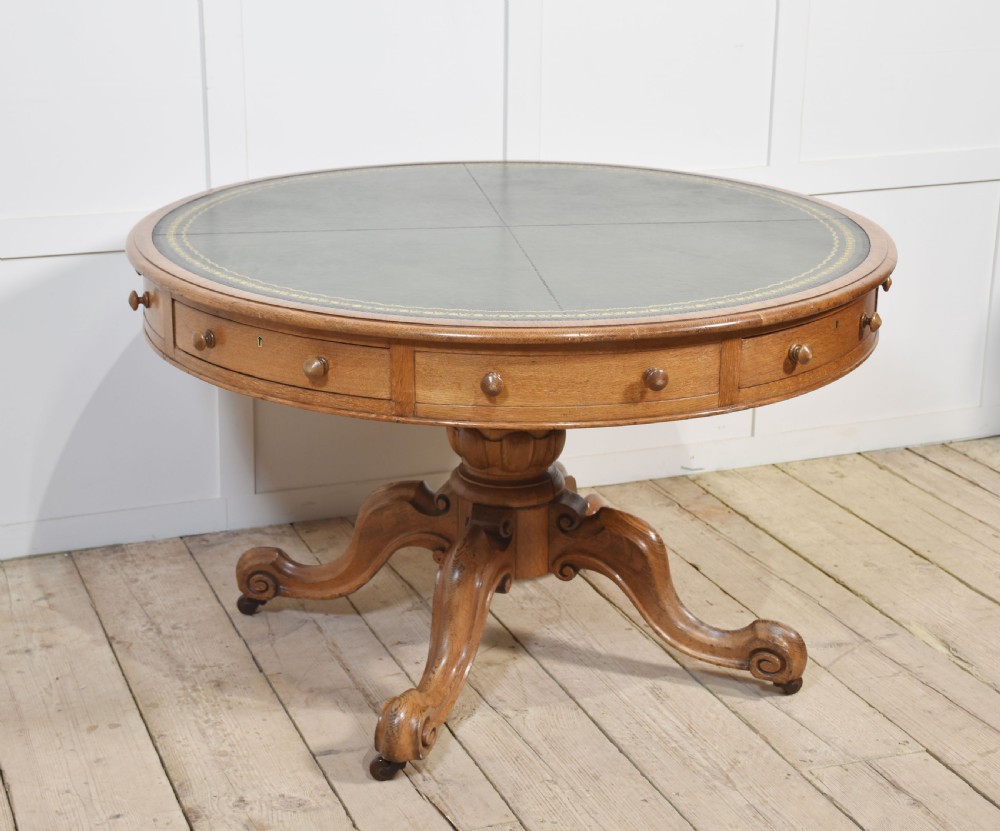 A 19th-century circular oak drum table featuring a green leather inset top with gilt tooling, multiple frieze drawers, and a turned pedestal on scroll legs with brass castors.