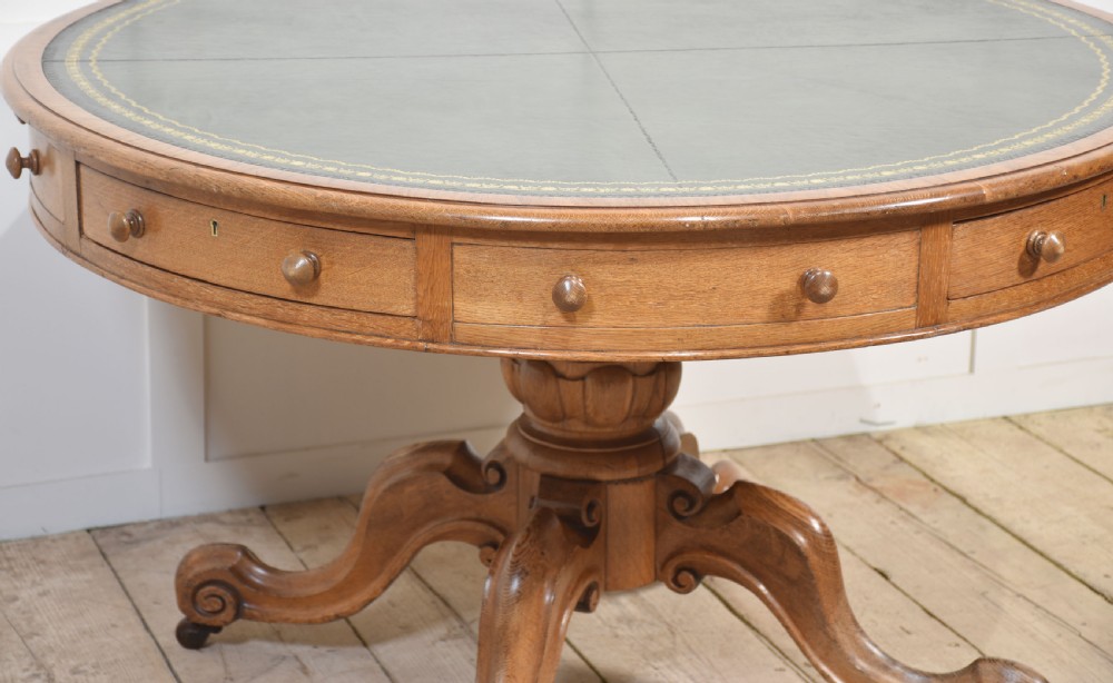 A 19th-century circular oak drum table featuring a green leather inset top with gilt tooling, multiple frieze drawers, and a turned pedestal on scroll legs with brass castors. View V