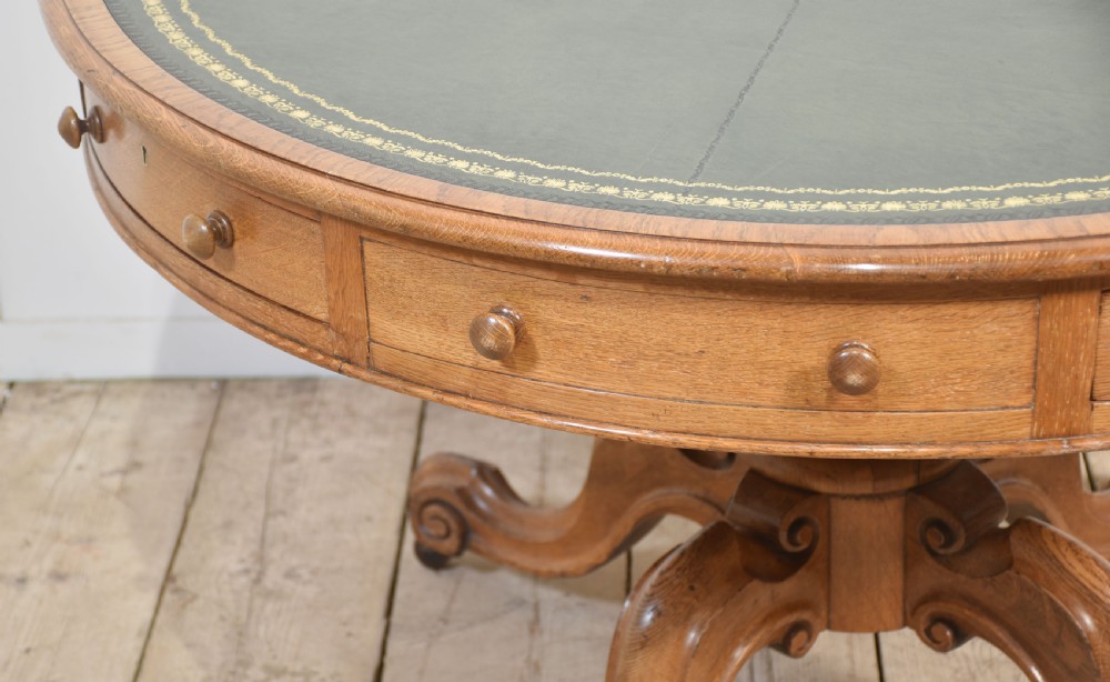 A 19th-century circular oak drum table featuring a green leather inset top with gilt tooling, multiple frieze drawers, and a turned pedestal on scroll legs with brass castors. View VIII