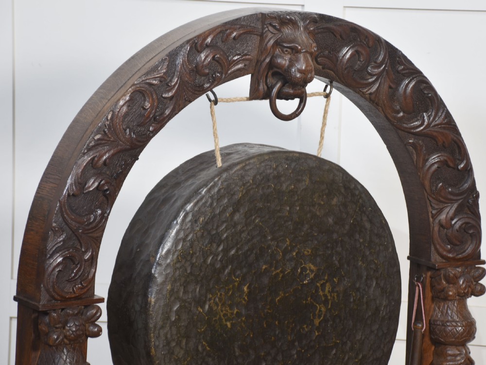 A dark carved oak arched frame featuring a central lion head mask housing a hammered circular brass gong and suspended striking mallet. View V