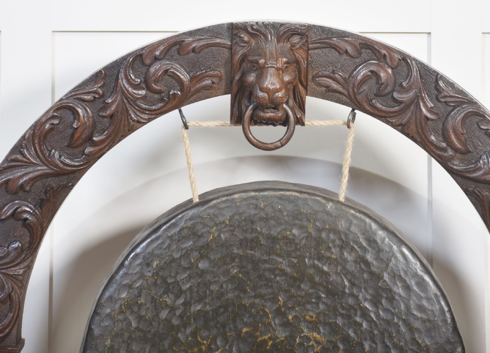 A dark carved oak arched frame featuring a central lion head mask housing a hammered circular brass gong and suspended striking mallet. View VII