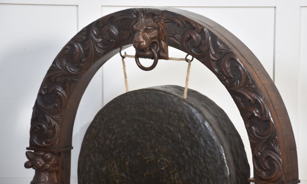 A dark carved oak arched frame featuring a central lion head mask housing a hammered circular brass gong and suspended striking mallet. View XI