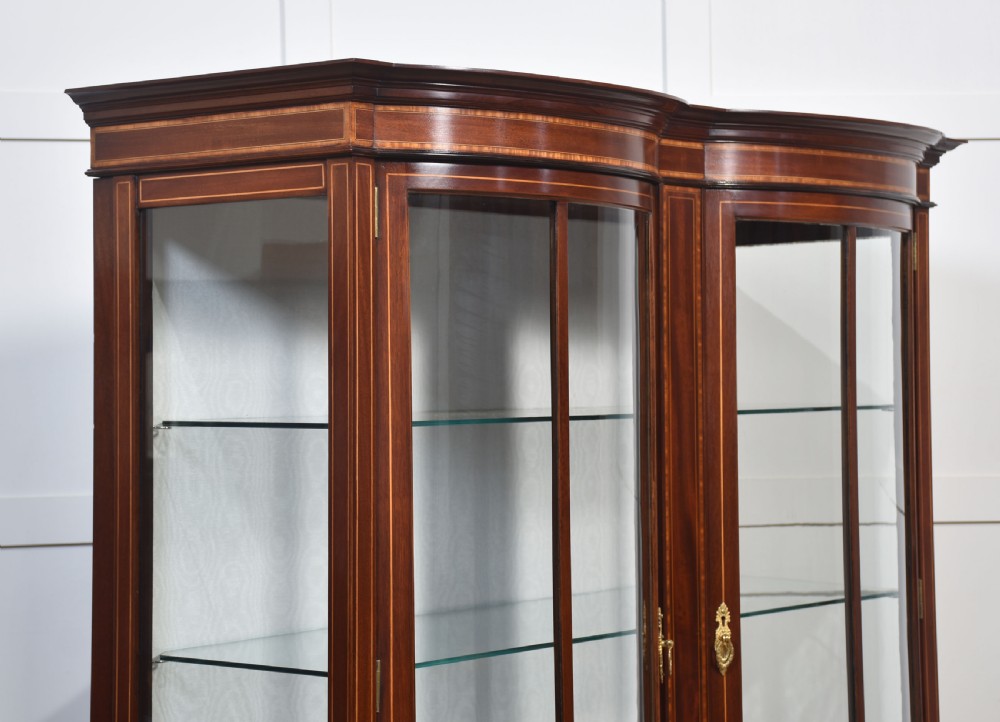 An Edwardian mahogany double bow-front display cabinet featuring glazed doors, fine satinwood inlay, glass shelves, and a lower shelf supported by tapered legs. View V