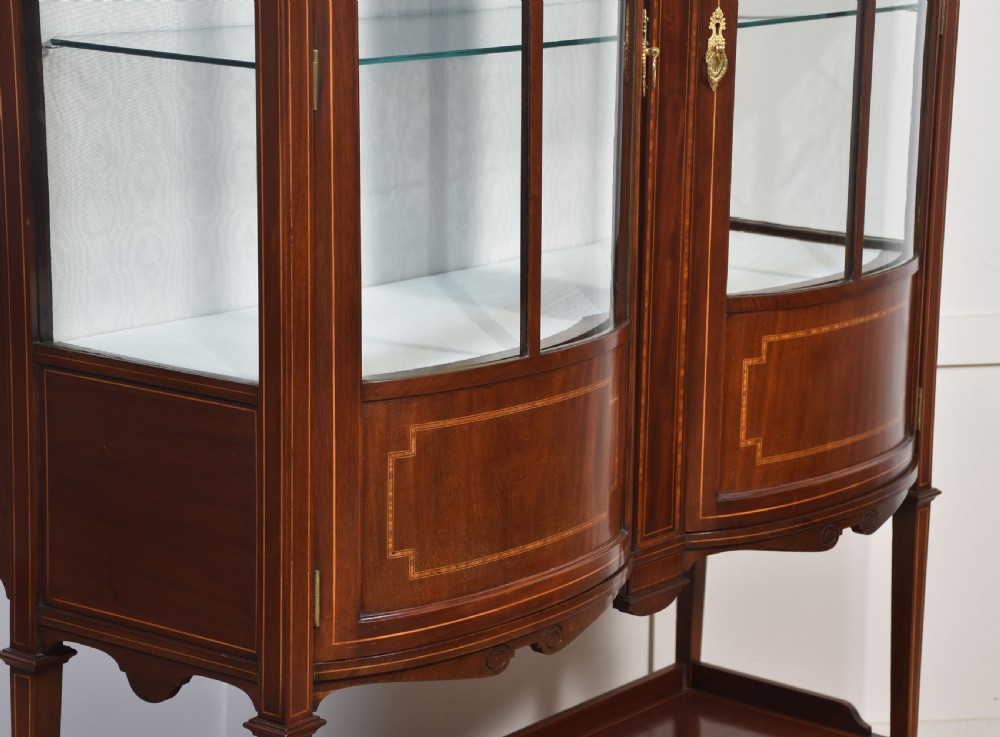 An Edwardian mahogany double bow-front display cabinet featuring glazed doors, fine satinwood inlay, glass shelves, and a lower shelf supported by tapered legs. View VII
