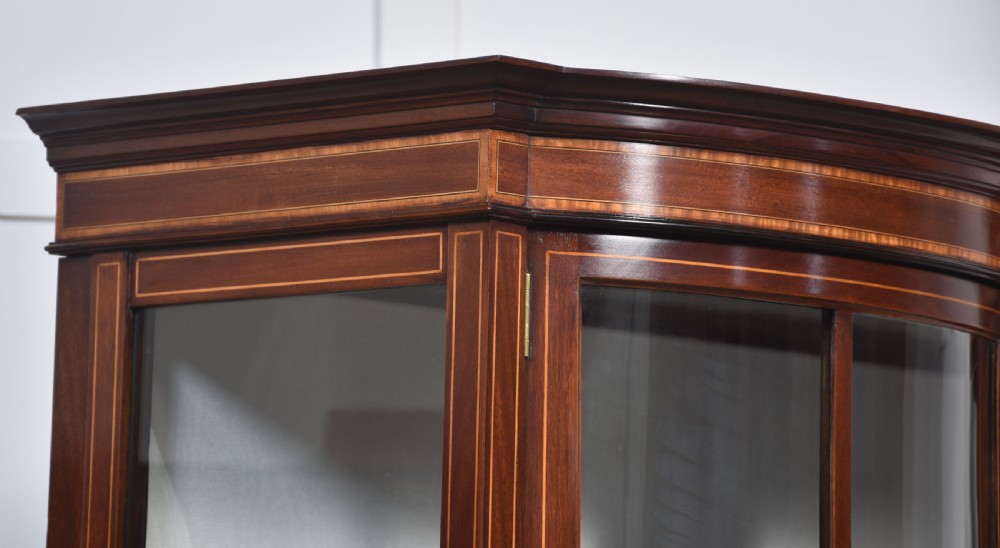 An Edwardian mahogany double bow-front display cabinet featuring glazed doors, fine satinwood inlay, glass shelves, and a lower shelf supported by tapered legs. View X