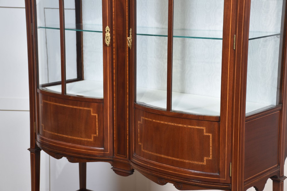 An Edwardian mahogany double bow-front display cabinet featuring glazed doors, fine satinwood inlay, glass shelves, and a lower shelf supported by tapered legs. View XII