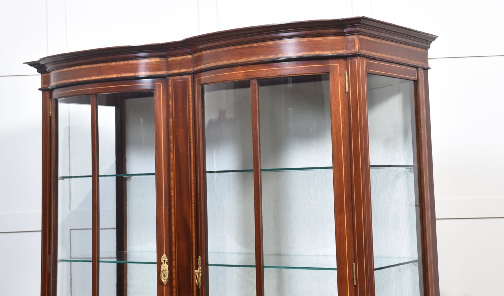 An Edwardian mahogany double bow-front display cabinet featuring glazed doors, fine satinwood inlay, glass shelves, and a lower shelf supported by tapered legs. View XIII
