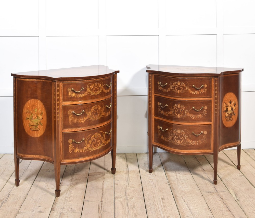 A near pair of bow-fronted mahogany chests of drawers featuring detailed floral marquetry inlay, brass handles, and slender tapered legs.