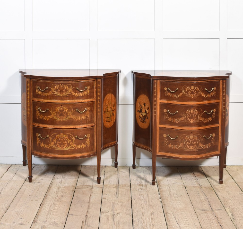 A near pair of bow-fronted mahogany chests of drawers featuring detailed floral marquetry inlay, brass handles, and slender tapered legs. View II