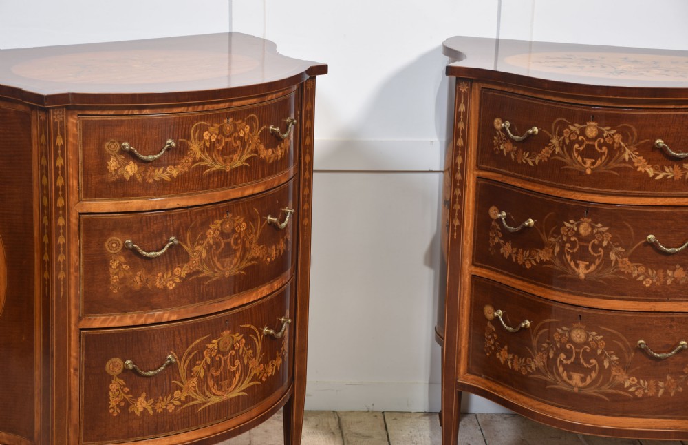 A near pair of bow-fronted mahogany chests of drawers featuring detailed floral marquetry inlay, brass handles, and slender tapered legs. View V