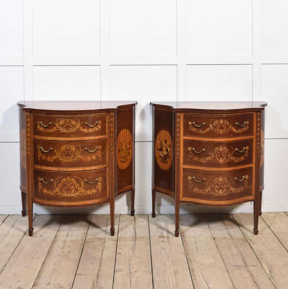 A near pair of bow-fronted mahogany chests of drawers featuring detailed floral marquetry inlay, brass handles, and slender tapered legs. View VI