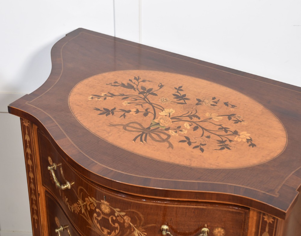A near pair of bow-fronted mahogany chests of drawers featuring detailed floral marquetry inlay, brass handles, and slender tapered legs. View IX