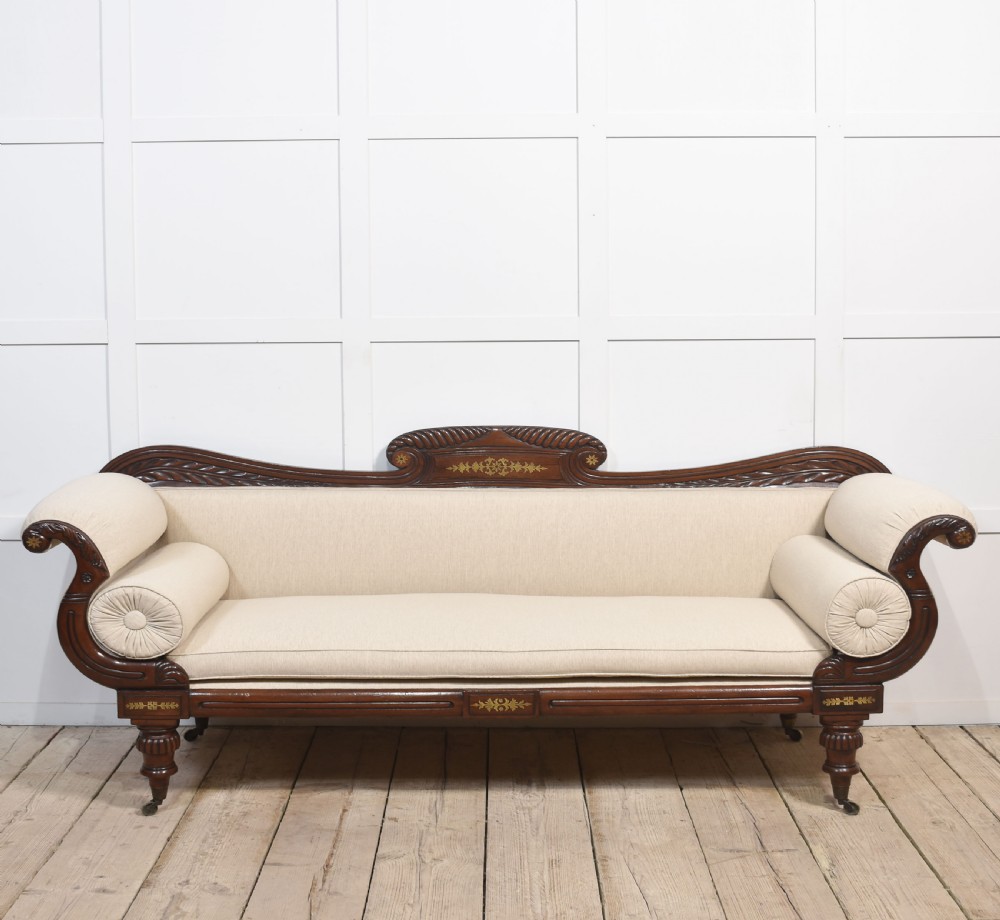 An antique Regency mahogany settee featuring brass inlay accents along the crest rail and apron, scrolled arms with matching bolster cushions, cream fabric upholstery, and turned legs on brass castors.