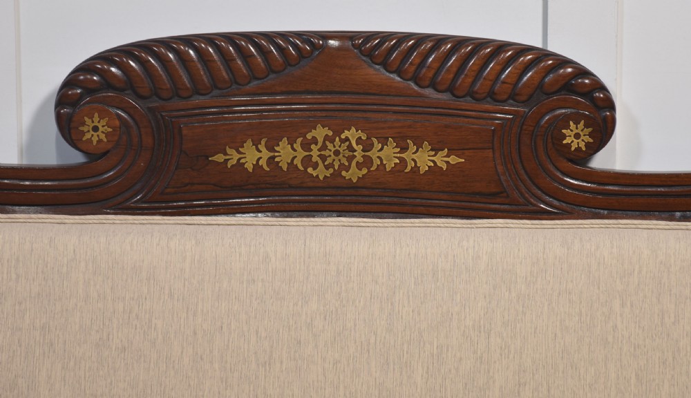 An antique Regency mahogany settee featuring brass inlay accents along the crest rail and apron, scrolled arms with matching bolster cushions, cream fabric upholstery, and turned legs on brass castors. View II