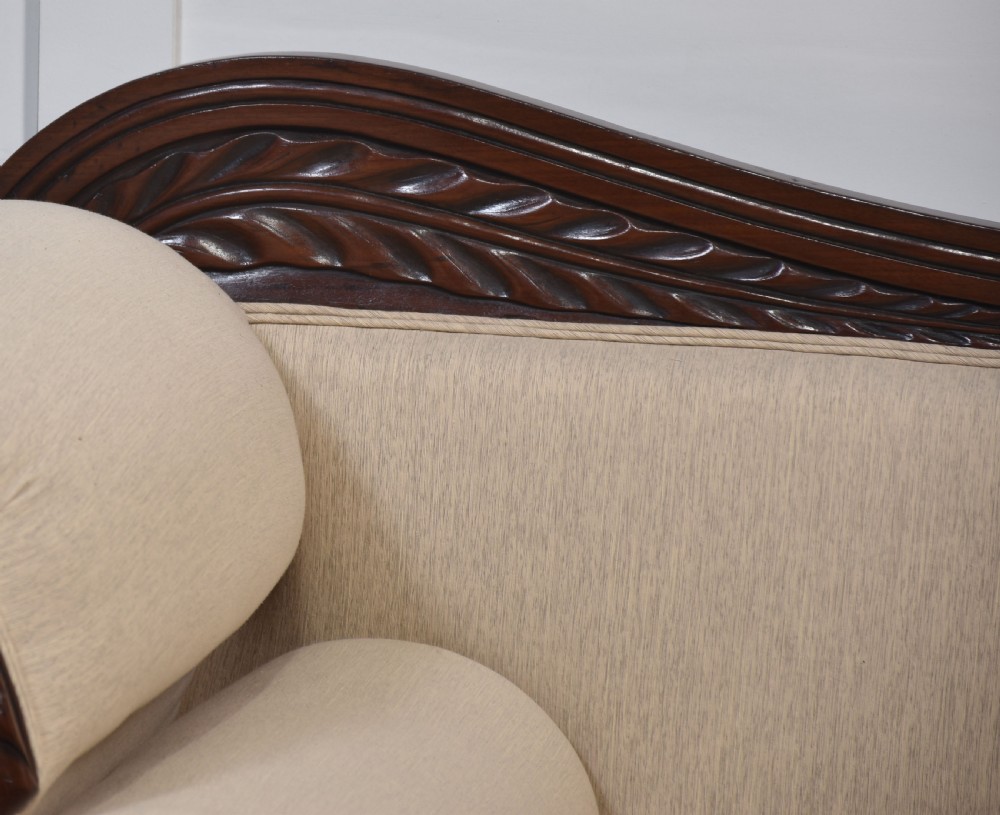 An antique Regency mahogany settee featuring brass inlay accents along the crest rail and apron, scrolled arms with matching bolster cushions, cream fabric upholstery, and turned legs on brass castors. View V
