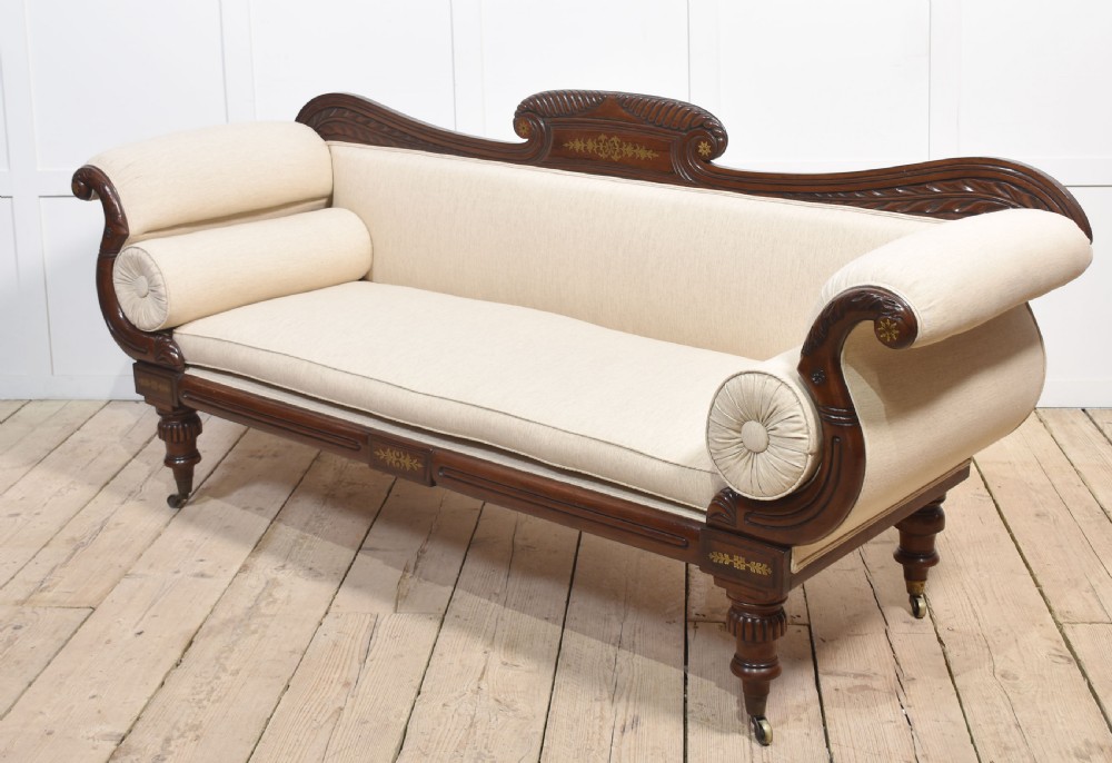 An antique Regency mahogany settee featuring brass inlay accents along the crest rail and apron, scrolled arms with matching bolster cushions, cream fabric upholstery, and turned legs on brass castors. View VI