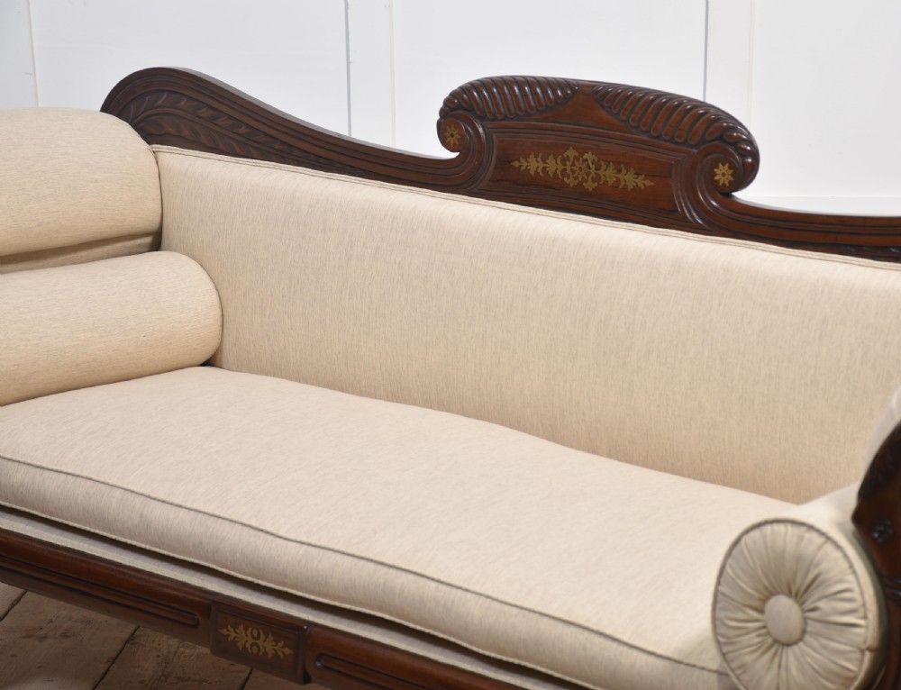 An antique Regency mahogany settee featuring brass inlay accents along the crest rail and apron, scrolled arms with matching bolster cushions, cream fabric upholstery, and turned legs on brass castors. View VII