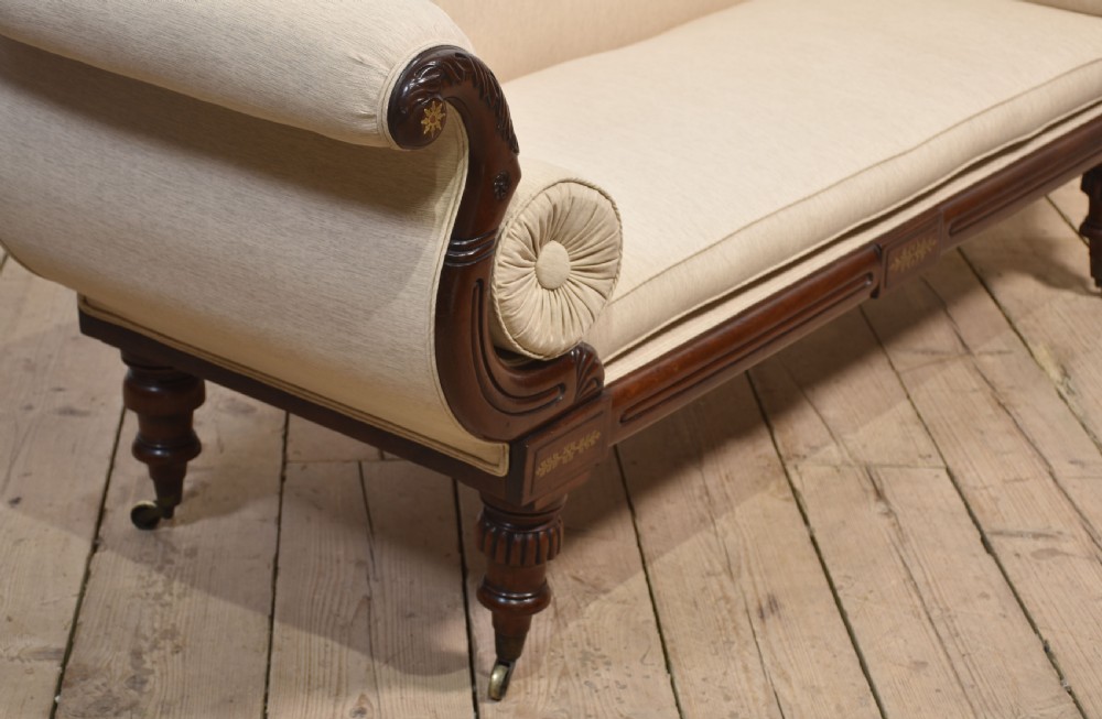 An antique Regency mahogany settee featuring brass inlay accents along the crest rail and apron, scrolled arms with matching bolster cushions, cream fabric upholstery, and turned legs on brass castors. View XI