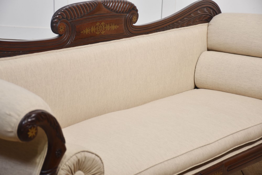 An antique Regency mahogany settee featuring brass inlay accents along the crest rail and apron, scrolled arms with matching bolster cushions, cream fabric upholstery, and turned legs on brass castors. View XIII
