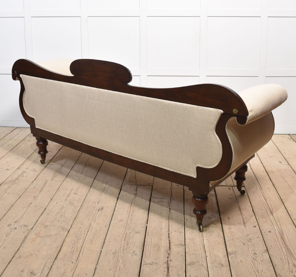 An antique Regency mahogany settee featuring brass inlay accents along the crest rail and apron, scrolled arms with matching bolster cushions, cream fabric upholstery, and turned legs on brass castors. View XIV