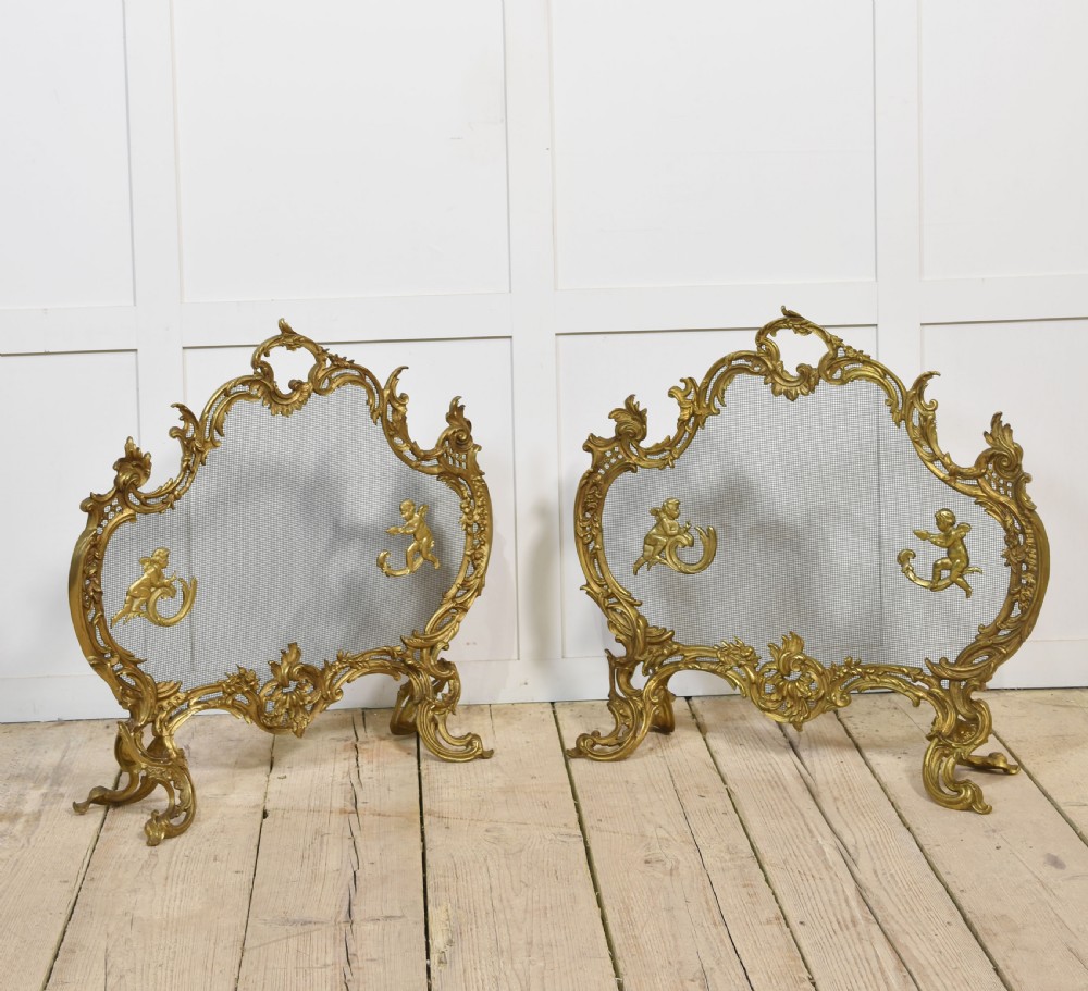 A pair of ornate Louis XV style gilt bronze fire screens featuring Rococo scrollwork, cherub mounts, and wire mesh panels on cabriole feet.