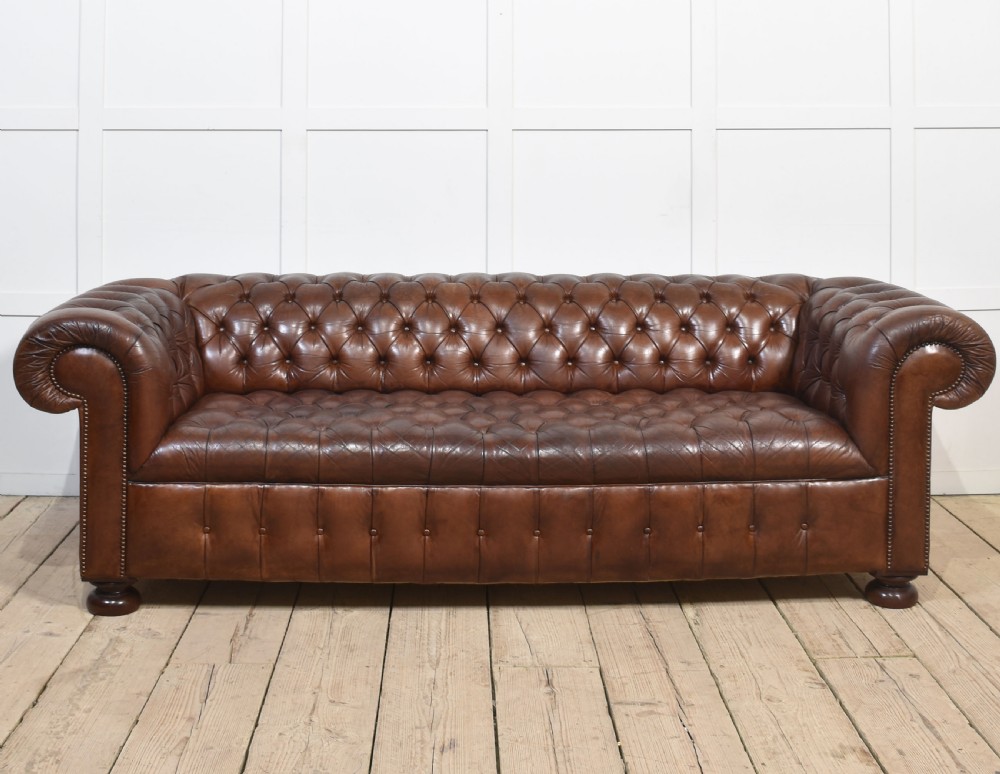 A three-seater brown leather Chesterfield sofa featuring deep buttoned upholstery, rolled arms, brass stud detailing, and turned wooden feet.