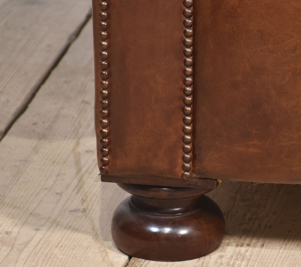 A three-seater brown leather Chesterfield sofa featuring deep buttoned upholstery, rolled arms, brass stud detailing, and turned wooden feet. View III