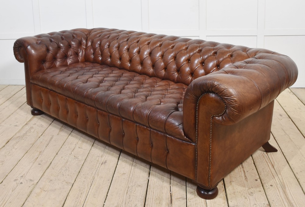 A three-seater brown leather Chesterfield sofa featuring deep buttoned upholstery, rolled arms, brass stud detailing, and turned wooden feet. View V