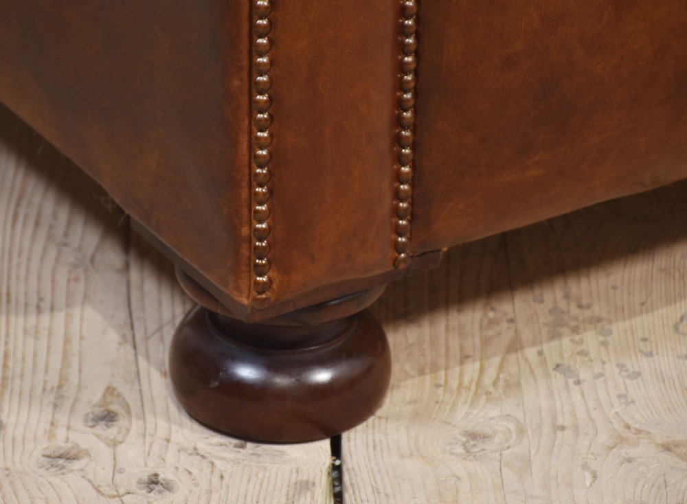 A three-seater brown leather Chesterfield sofa featuring deep buttoned upholstery, rolled arms, brass stud detailing, and turned wooden feet. View VI