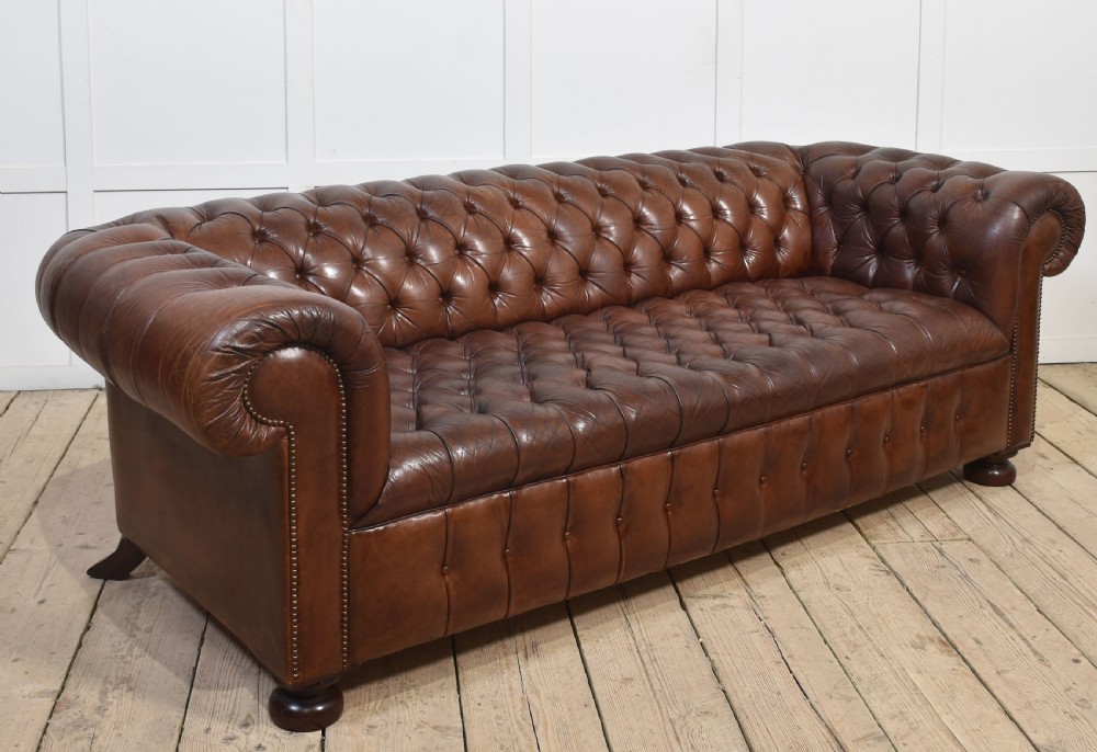 A three-seater brown leather Chesterfield sofa featuring deep buttoned upholstery, rolled arms, brass stud detailing, and turned wooden feet. View VIII