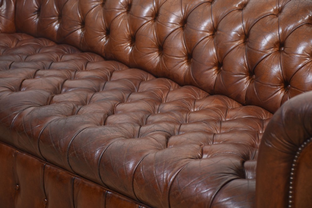 A three-seater brown leather Chesterfield sofa featuring deep buttoned upholstery, rolled arms, brass stud detailing, and turned wooden feet. View IX