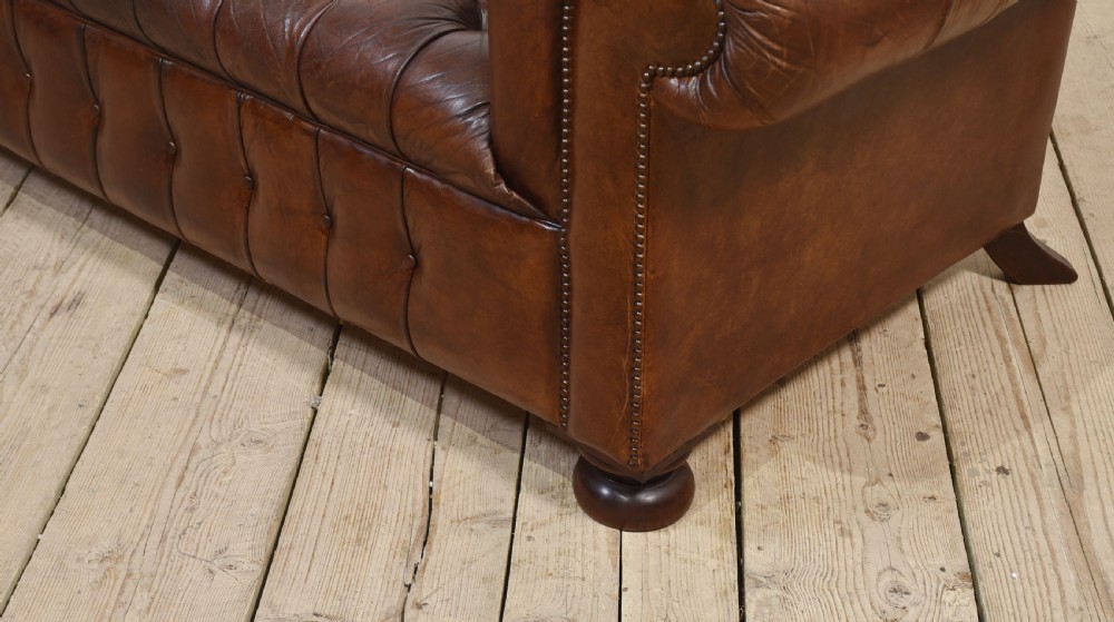 A three-seater brown leather Chesterfield sofa featuring deep buttoned upholstery, rolled arms, brass stud detailing, and turned wooden feet. View X
