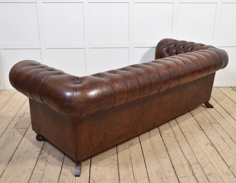 A three-seater brown leather Chesterfield sofa featuring deep buttoned upholstery, rolled arms, brass stud detailing, and turned wooden feet. View XI