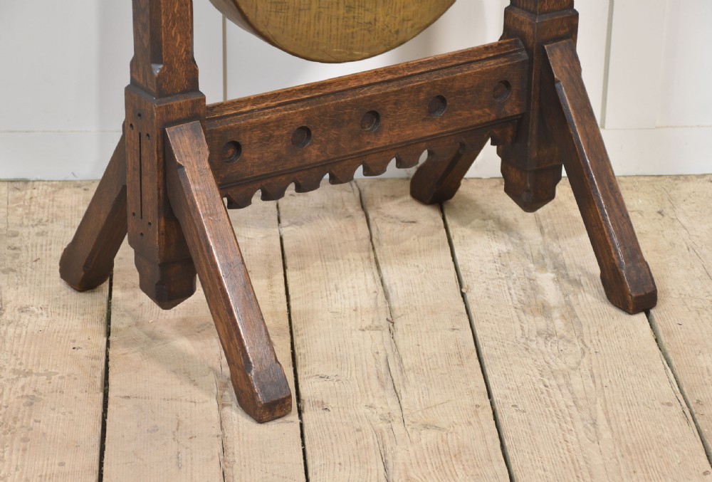 A Gothic Revival dark oak floor-standing frame featuring carved trefoil piercings and holding a circular hand-hammered brass dinner gong with its original striker. View II