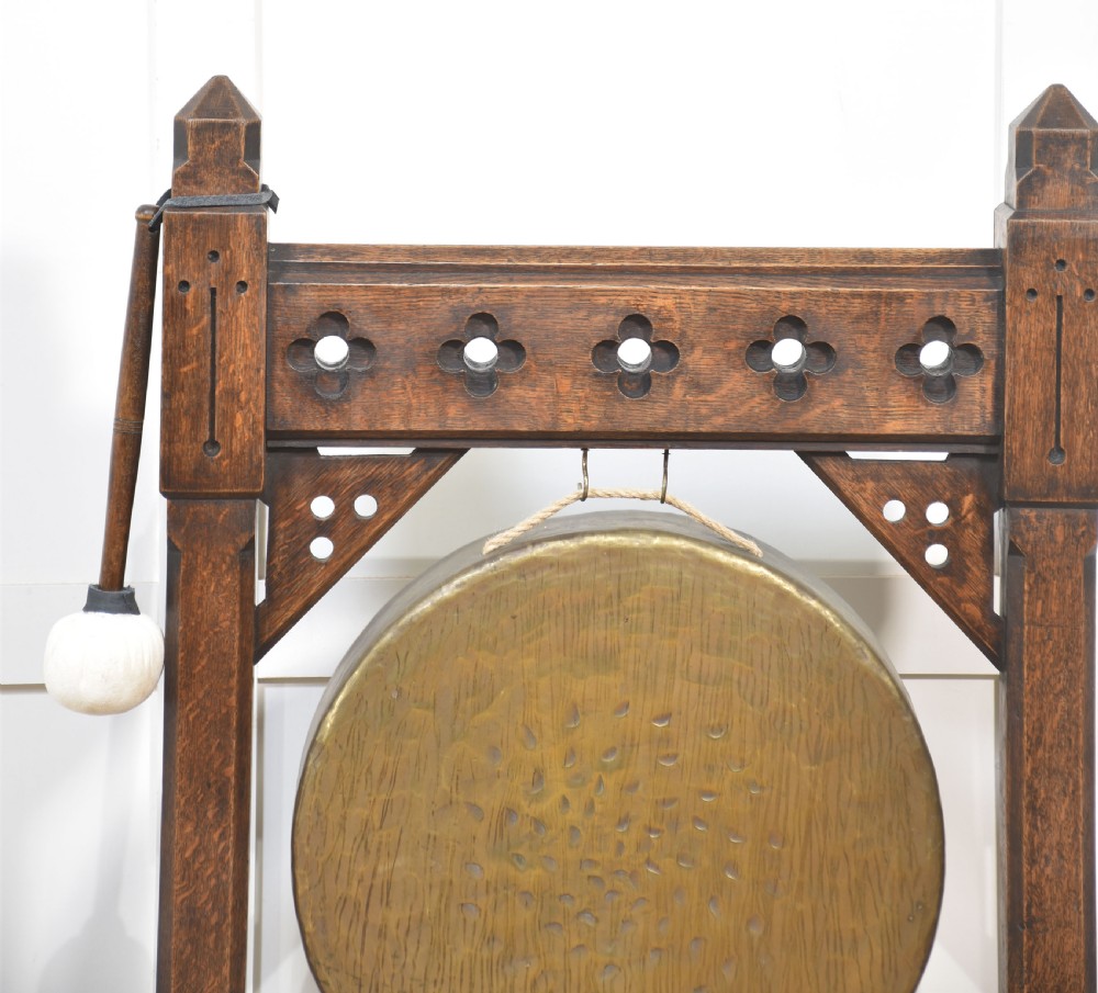 A Gothic Revival dark oak floor-standing frame featuring carved trefoil piercings and holding a circular hand-hammered brass dinner gong with its original striker. View V