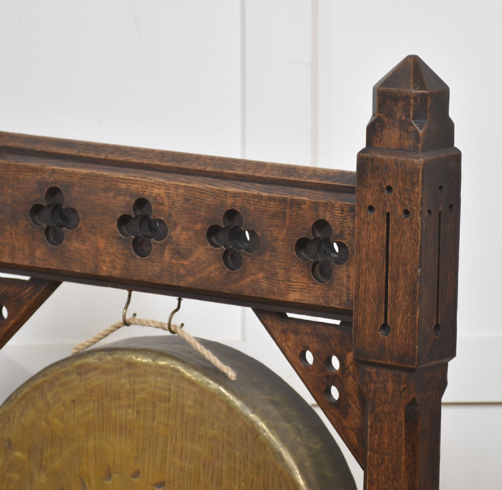 A Gothic Revival dark oak floor-standing frame featuring carved trefoil piercings and holding a circular hand-hammered brass dinner gong with its original striker. View VI