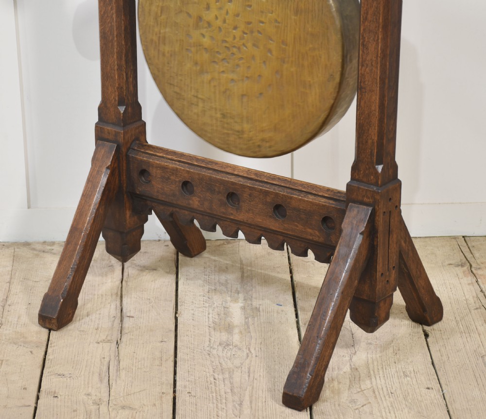 A Gothic Revival dark oak floor-standing frame featuring carved trefoil piercings and holding a circular hand-hammered brass dinner gong with its original striker. View IX