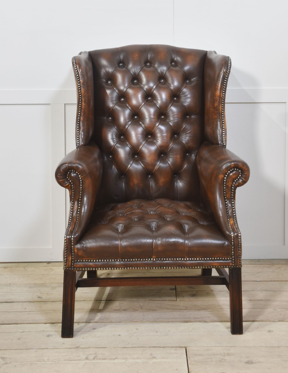A warm brown leather wingback armchair with deep buttoned tufting, brass stud detailing, and dark wooden legs.