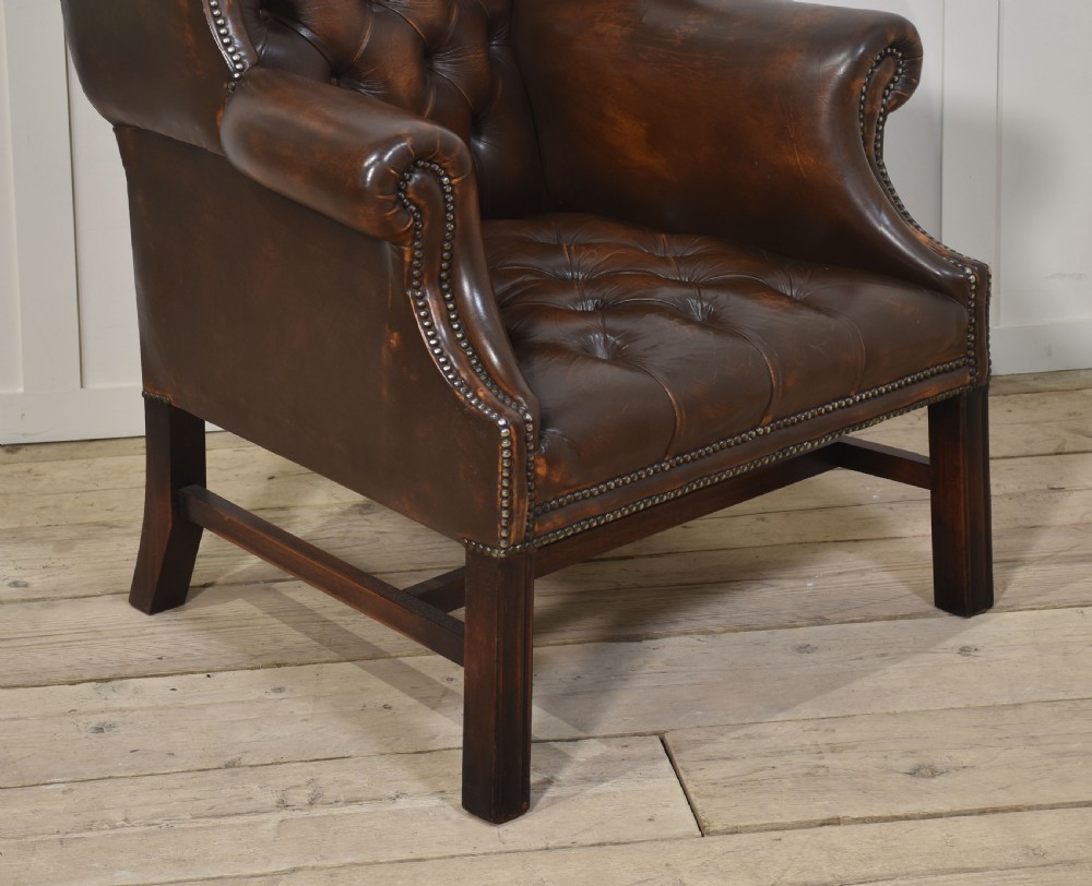 A warm brown leather wingback armchair with deep buttoned tufting, brass stud detailing, and dark wooden legs. View VII