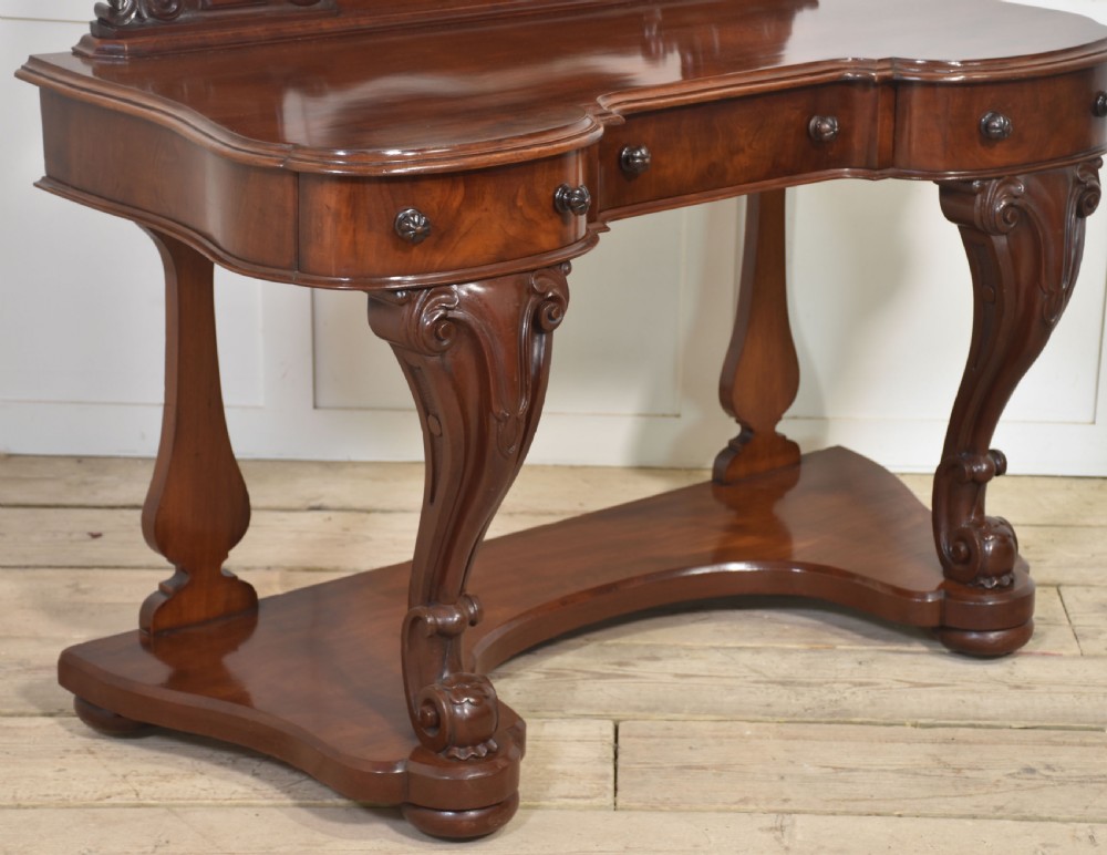 A Victorian mahogany duchess dressing table featuring an arch-topped swivel mirror flanked by carved uprights above a serpentine front desk with drawers and a shaped platform base. View XIV